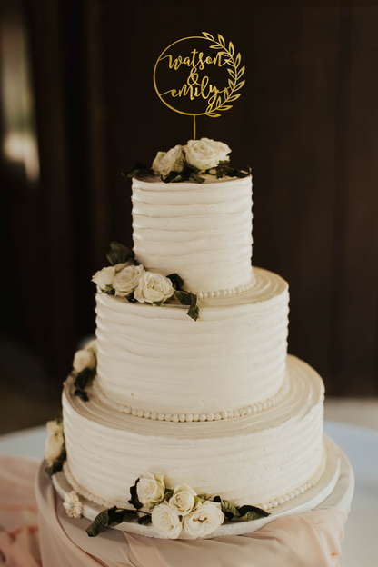 calligraphy personalized golden wreath style cake topper made of wood on top of the white simple wedding cake. 